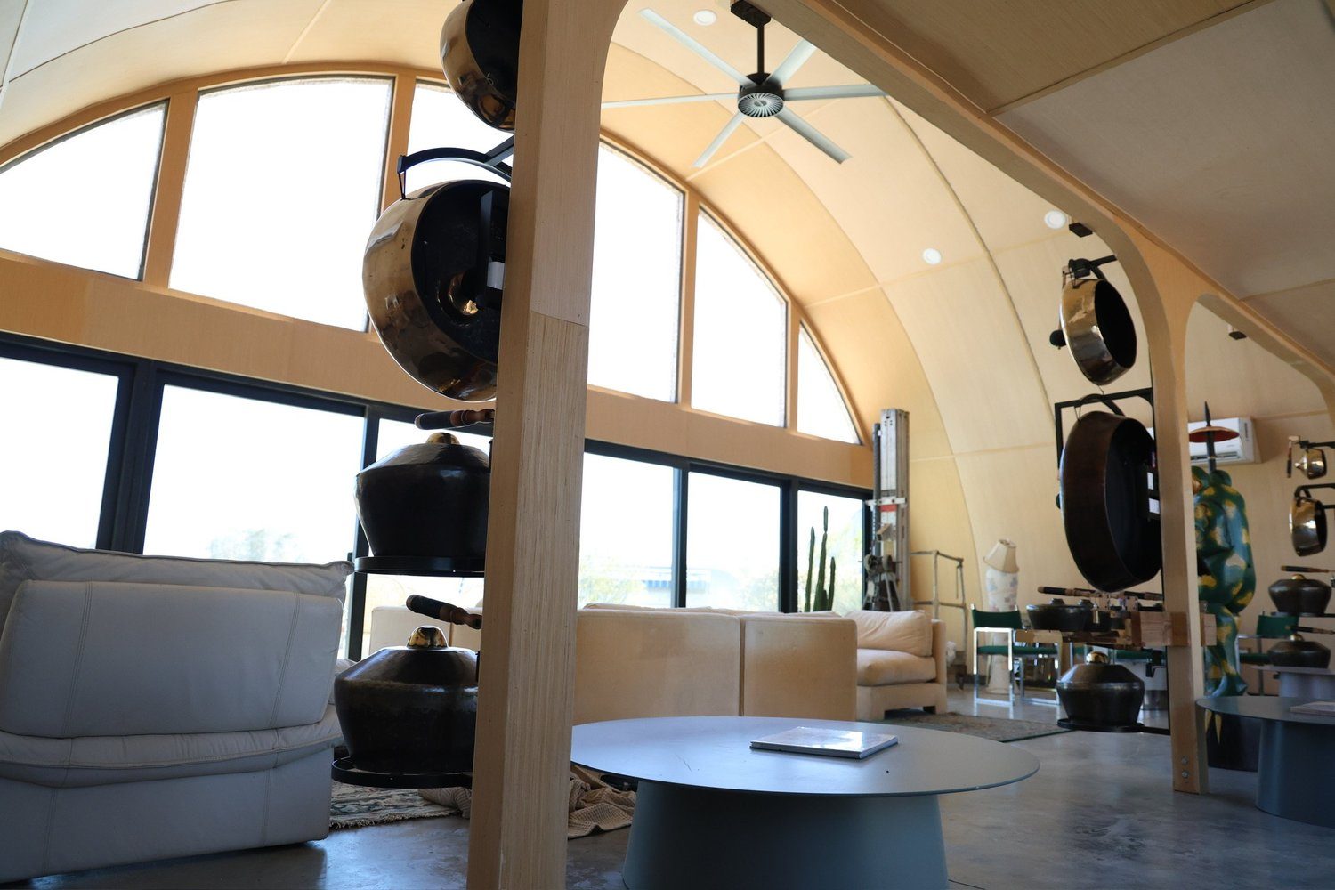 Inside the vaulted Center, bronze kettle gongs beside soft cream seating under a sweeping arched window