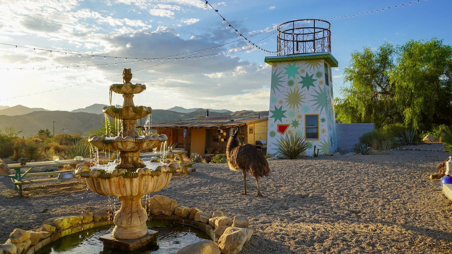 An emu wandering past the tiered stone fountain toward the star painted tower, string lights and golden mountains behind
