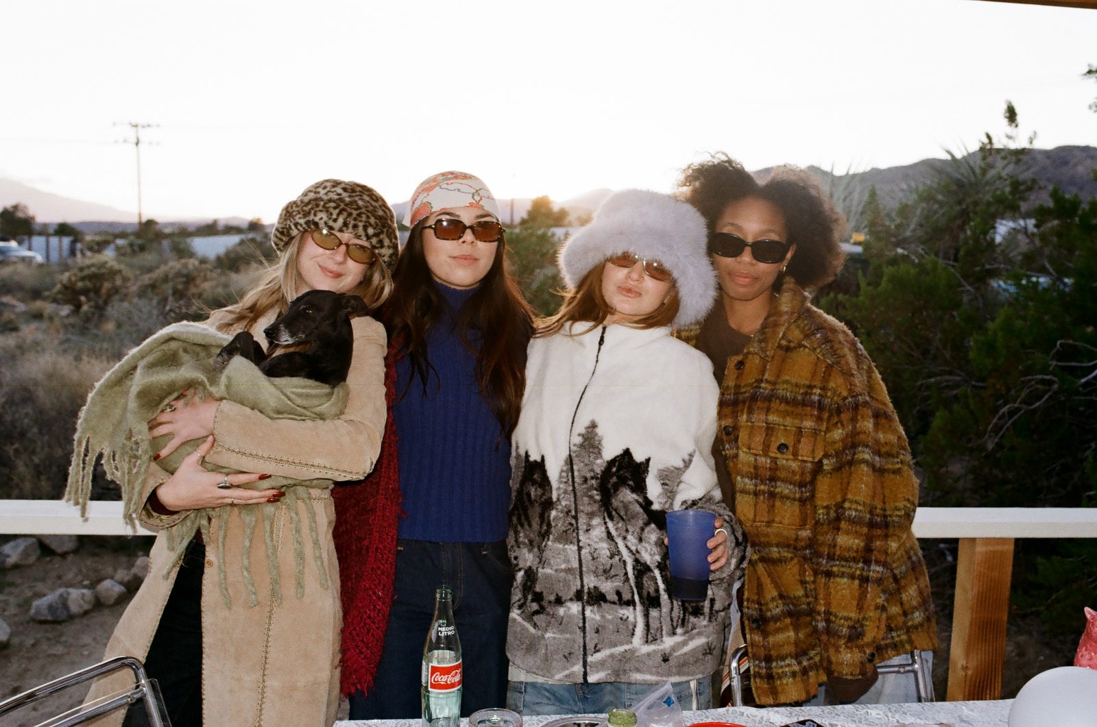 Four friends bundled in coats and sunglasses on a desert deck at golden hour, one cradling a small dog in her arms