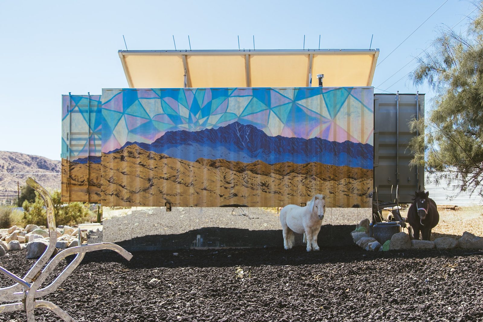 Woodamon’s mountain mural head on across the full length of the container, a violet range under a faceted turquoise sky, a white mini pony standing in front of it